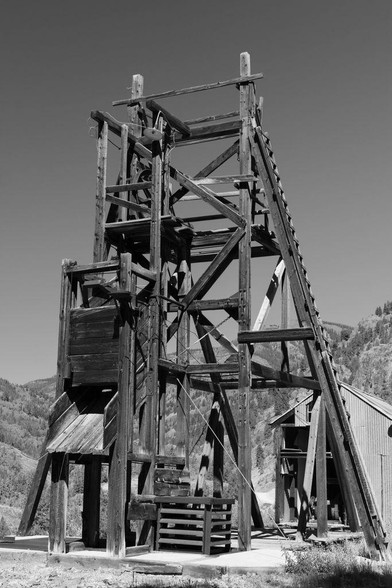 Black and white photo of a historic wooden mine tower structure set against a mountainous backdrop near Lake City, Colorado. The rugged beams and pulley system reflect early mining engineering.