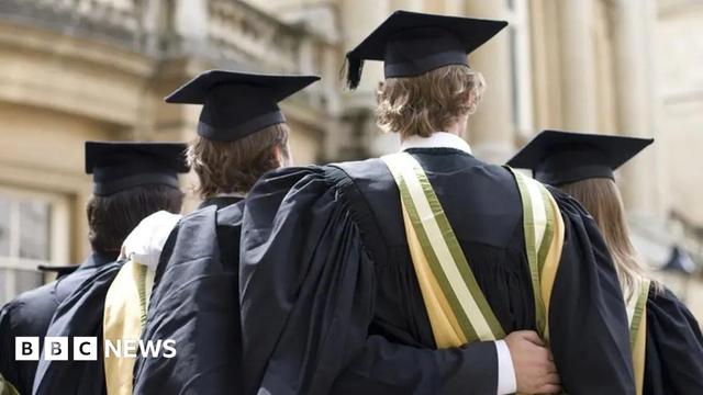 Four students wearing black graduation gowns and caps with yellow and green sashes stand closely together with their backs to the camera. One student has an arm around another’s shoulder. They are outdoors in front of a historic building with ornate architecture.