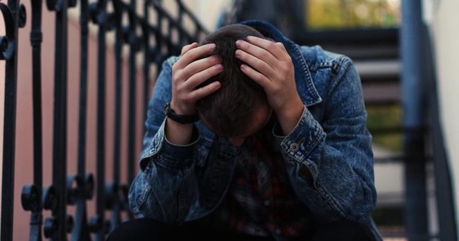 Sad young boy in jeans jacket with his hands on the head is sitting on city stair.