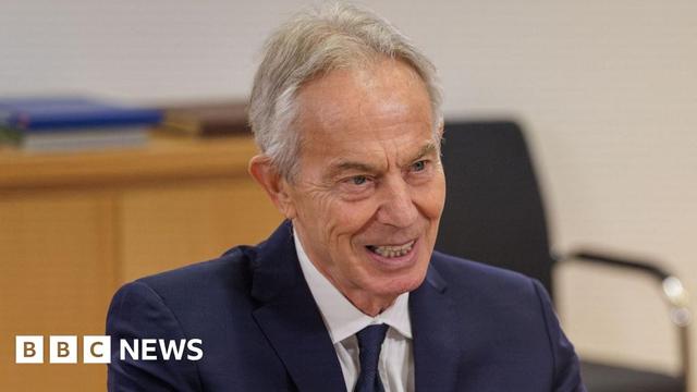 Tony Blair wears a blue suit and tie with a white shirt. He has grey hair and there is a wooden desk behind him.