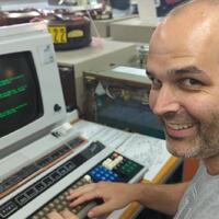 Photo of me, a white man with a short dark beard, and dark hair, smiling at the camera, while sitting in front of a vintage terminal with green text on the screen.
