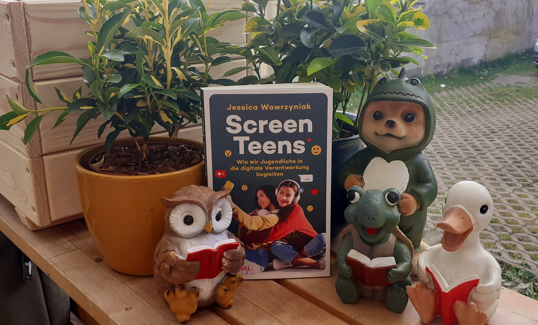 Das Buch "Screen Teens" steht auf einer Bank. Dahinter grüne Pflanzen. Davor Gartenfiguren, die ein Buch lesen: Eine Eule, eine Schildkröte und eine Gans