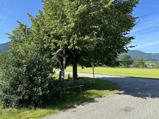 Bank im Schatten vor einem Wegkreuz, daneben lehnt ein Fahrrad an einem viel Schatten spendenden Baum, ein Weg mit schon rissigem Asphalt, im Hintergrund Berge