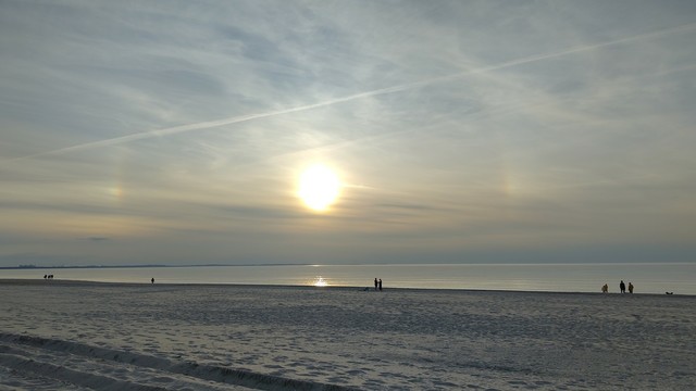 Ostsee-Strand, glattes Wasser, leichte Bewölkung, ein kleines Halo im Himmel.