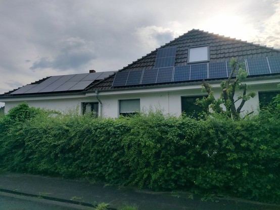 Seitenansicht eines Wohnhauses mit einer Hecke davor, auf dem Dach mehrere Solarmodule und ein Dachfenster