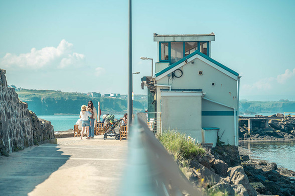 Eine Hafenlandschaft. Auf der linken Seite sommerlich gekleidete Menschen in der Sonne. Auf der rechten Seite ein Café, das von der Seite wie ein Bootshaus aussieht.
Drumherum erkennt man etwas Meer und blauen Himmel.
Getrennt wird das Bild in der Bildmitte durch ein verschwommenes Geländer und einen anschließenden Laternenmast.