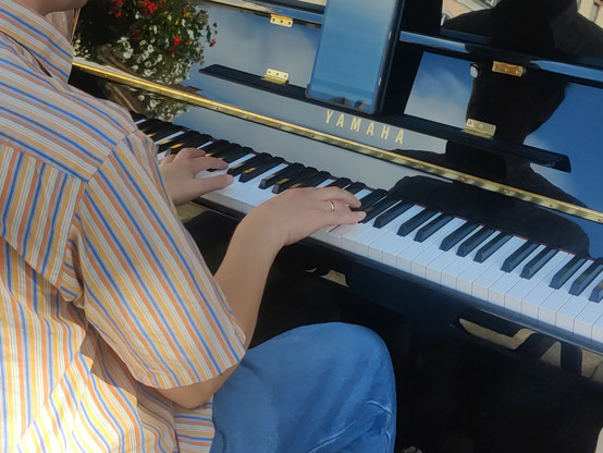 A person playing a shiny black upright piano, a colourful striped shirt, the right leg in blue jeans, and the hands are visible, shot from the rear right. A tablet sits on the note stand, flowers and blue sky are reflected in the piano.