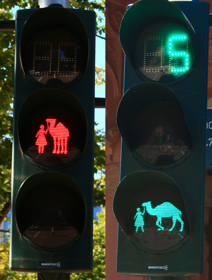 Zwei engzugeschnittene Bilder einer Fußgängerampel, links rot zeigend, mit beleuchtetem Laub im Hintergrund, rechts grün zeigend, mit einem Gebäude im Hintergrund. Die Figuren zeigen kein traditionelles Ampelmännchen, sondern eine stilisierte Person mit ausladendem Kleid, die ein Kamel an der Leine führt, stehend für rot, und gehend für grün. Aufer der grün zeigenden Ampel ist oben ein 