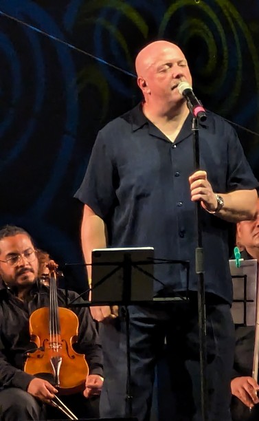 Die Aufnahme zeigt Ronan Harris stehend als Ganzkörperportrait auf der Konzertbühne umgeben von Musikern der Philharmie Leipzig. (leider nur Handyqualität) 