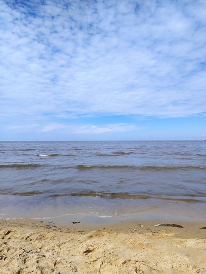 Sanfte Wellen rollen an den Sandstrand. Obere Bildhälfte Himmel mit leichter Bewölkung, dann braun-blaues Meer, unterstes Viertel heller Sandstrand.