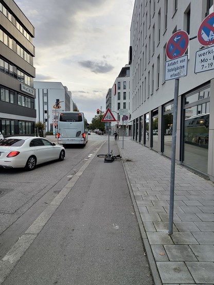 Zeichen 138-10, Achtung Radverkehr, auf einem mobilen Pfosten mittig(!) auf einem Radweg. An den Pfosten gammelt eine Fahrradleiche vor sich hin.