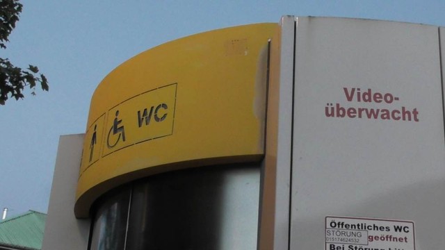 Das Bild zeigt einen Ausschnitt eines öffentlichen WC-Gebäudes. Oben befindet sich ein gelbes Schild mit den Symbolen für ein Unisex-WC und ein behindertengerechtes WC, daneben steht „WC“. Auf der rechten Seite des Gebäudes steht in roter Schrift „Video überwacht“. Darunter befindet sich ein kleineres Schild mit der Aufschrift „Öffentliches WC“, auf dem Kontaktdaten für den Fall einer Störung zu finden sind.

Foto (c) 2024 Claus Schönleber