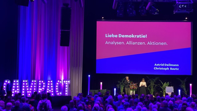 Eine Bühne ist zu sehen, auf der eine Präsentation mit der Überschrift „Liebe Demokratie! Analysen. Allianzen. Aktionen.“ angezeigt wird. Auf der rechten Seite des Bildschirms stehen die Namen „Astrid Deilmann“ und „Christoph Bautz“. Der Hintergrund der Bühne ist in blauem und pinkem Licht beleuchtet. Links ist der Schriftzug „CAMPACT“ in großen, beleuchteten Buchstaben sichtbar. Vor der Bühne befindet sich eine sitzende Zuschauergruppe, während auf der Bühne zwei Personen zu sehen sind, eine Frau in heller Bluse und ein Mann, der spricht. Pflanzen und Blumenarrangements sind ebenfalls auf der Bühne platziert.