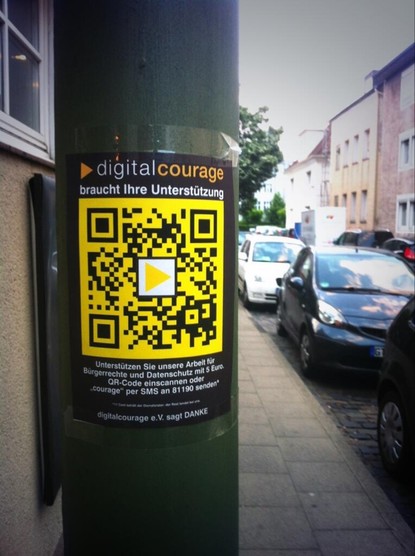 Ein Aufkleber, das an einem grünen Laternenpfahl befestigt ist. Der obere Teil zeigt das Logo von digitalcourage und den Text: „braucht Ihre Unterstützung“. Darunter ist ein großer gelber QR-Code mit dem Digitalcourage-Logo in der Mitte abgebildet.

Im unteren Bereich steht:
„Unterstützen Sie unsere Arbeit für Bürgerrechte und Datenschutz mit 5 Euro. QR-Code einscannen oder ‘courage’ per SMS an 81190 senden.*“

Kleine Fußnote:
„*17 Cent bekommt der Dienstleister, der Rest kommt bei uns an.“

Ganz unten steht: „Digitalcourage e.V. sagt DANKE“.
