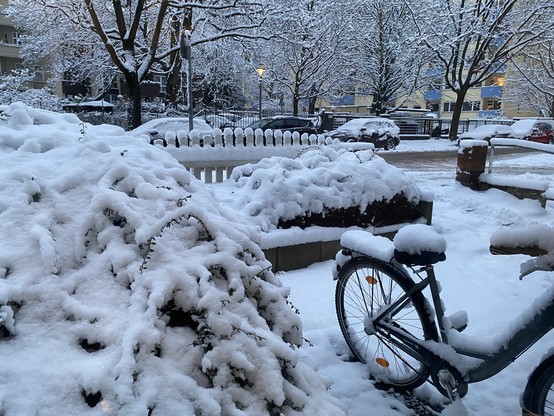 Ein verschneites Stadtbild in einem Wohngebiet: Büsche, ein Fahrrad und eine Bank sind dick mit Schnee bedeckt. Im Hintergrund stehen schneebedeckte Bäume, parkende Autos und ein mehrstöckiges Wohnhaus mit beleuchteten Fenstern. Eine Laterne spendet warmes Licht in der Dämmerung. Die Straßen sind teils geräumt, aber noch mit Schnee bedeckt.