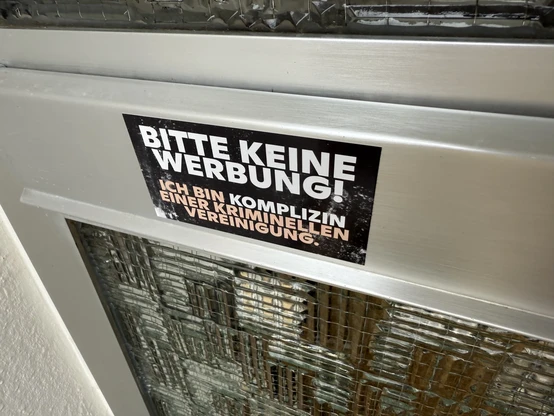 Aufkleber auf silbernem Briefschlitzdeckel in schwarz-weiß-rosa mit der Aufschrift "Bitte keine Werbung! Ich bin Komplizin einer kriminellen Vereinigung." von Zentrum für Politische Schönheit