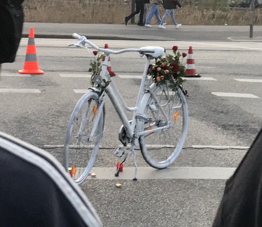 In der Mitte des Bildes ein weiß angestrichenes Damenrad, das auf einem Fußgängerüberweg steht, auf dem Gepäckträger wurden Blumen abgelegt. Um das Fahrrad herum wurden auf dem Asphalt Kerzen aufgestellt.
