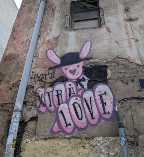 Das Bild zeigt eine Wand in einer Stadt, die mit Graffiti verziert ist. Die Wand ist aus verwittertem Beton und zeigt Spuren von Alter und Vernachlässigung, wie z. B. abblätterndes Putz und Risse. Auf der Wand ist ein buntes Graffiti zu sehen, das einen pinken, cartoonartigen Hasen mit einem schwarzen Hut und einem schwarzen Anzug darstellt. Der Hasen hat große Ohren und ein Herz auf der Nase. Neben dem Hasen steht in großen, pinken Buchstaben mit schwarzer Umrandung die Aufschrift "XIRÁ LOVE". Darunter ist in kleinerer Schrift "quarbi" zu lesen. Die Wand hat einen schmalen, dunklen Fensterbalken und ein graues Rohr, das an der Wand entlangläuft. Auf dem Rohr sind mehrere Klebeelemente zu sehen, darunter ein gelbes Etikett mit der Aufschrift "SUPER".