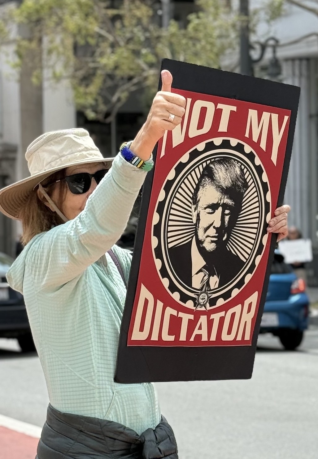 Das Bild zeigt eine Person mit Sonnenhut und Sonnenbrille, die ein Schild hochhĂ€lt. Auf dem Schild ist ein stilisiertes Schwarz-WeiĂ-PortrĂ€t von Donald Trump zu sehen. DarĂŒber steht in groĂen Buchstaben: âNOT MY DICTATORâ. Die Person zeigt zusĂ€tzlich den Daumen nach oben. Die Szene spielt im Freien. Das Bild zeigt eine Person mit Sonnenhut und Sonnenbrille, die ein Schild hochhĂ€lt. Auf dem Schild ist ein stilisiertes Schwarz-WeiĂ-PortrĂ€t von Donald Trump zu sehen. DarĂŒber steht in groĂen Buchstaben: âNOT MY DICTATORâ. Die Person zeigt zusĂ€tzlich den Daumen nach oben. Die Szene spielt im Freien.