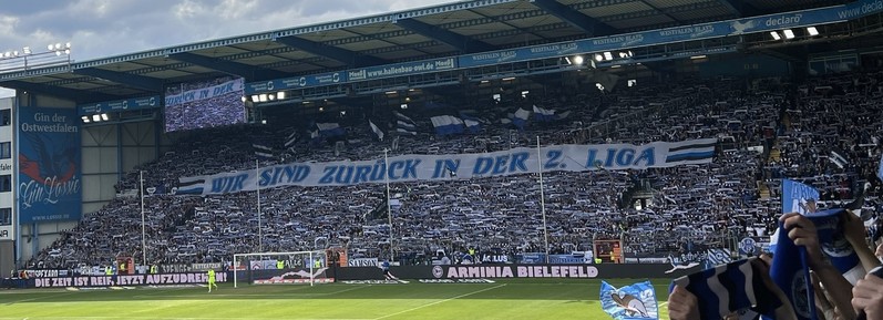 Das Bild zeigt die beeindruckende Choreografie der Fans von Arminia Bielefeld im heimischen Stadion anlässlich des Aufstiegs in die 2. Bundesliga.

Im Mittelpunkt steht ein großes Banner mit der Aufschrift “WIR SIND ZURÜCK IN DER 2. LIGA”, das von den Fans in der voll besetzten Tribüne gehalten wird. Die Fans tragen überwiegend blau-weiße Fanartikel wie Schals und Fahnen, passend zu den Vereinsfarben von Arminia Bielefeld.

Die Stimmung ist ausgelassen und jubelnd. Vor der Tribüne ist ein weiteres Banner zu sehen, das verkündet: “DIE ZEIT IST REIF, JETZT AUFZUDREHEN!”.

Das Stadion ist die Bielefelder Alm (das auch auf den gekauften Titel SchücoArena hört), wie an der Werbebande und dem Stadionumfeld erkennbar ist. Die Atmosphäre im Bild vermittelt Freude und Stolz über die Rückkehr in die zweite Liga.

Bild  Rena Tangens, cc by 4.0: