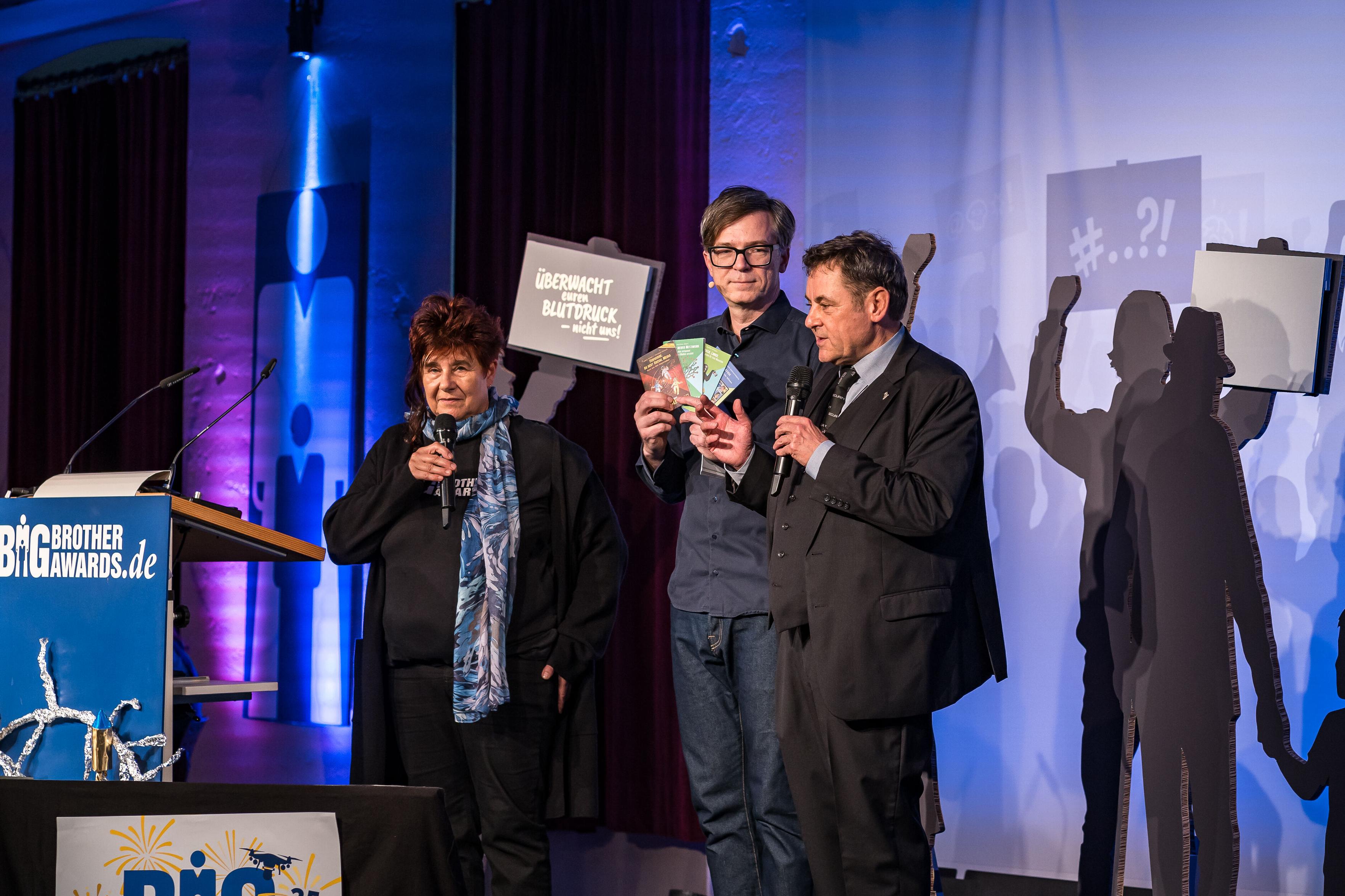 Ralph Caspers mit Reena Tangens und Padeluun auf der Bühne. Ralph Caspers hält mehrere Kurz&Mündig hoch.