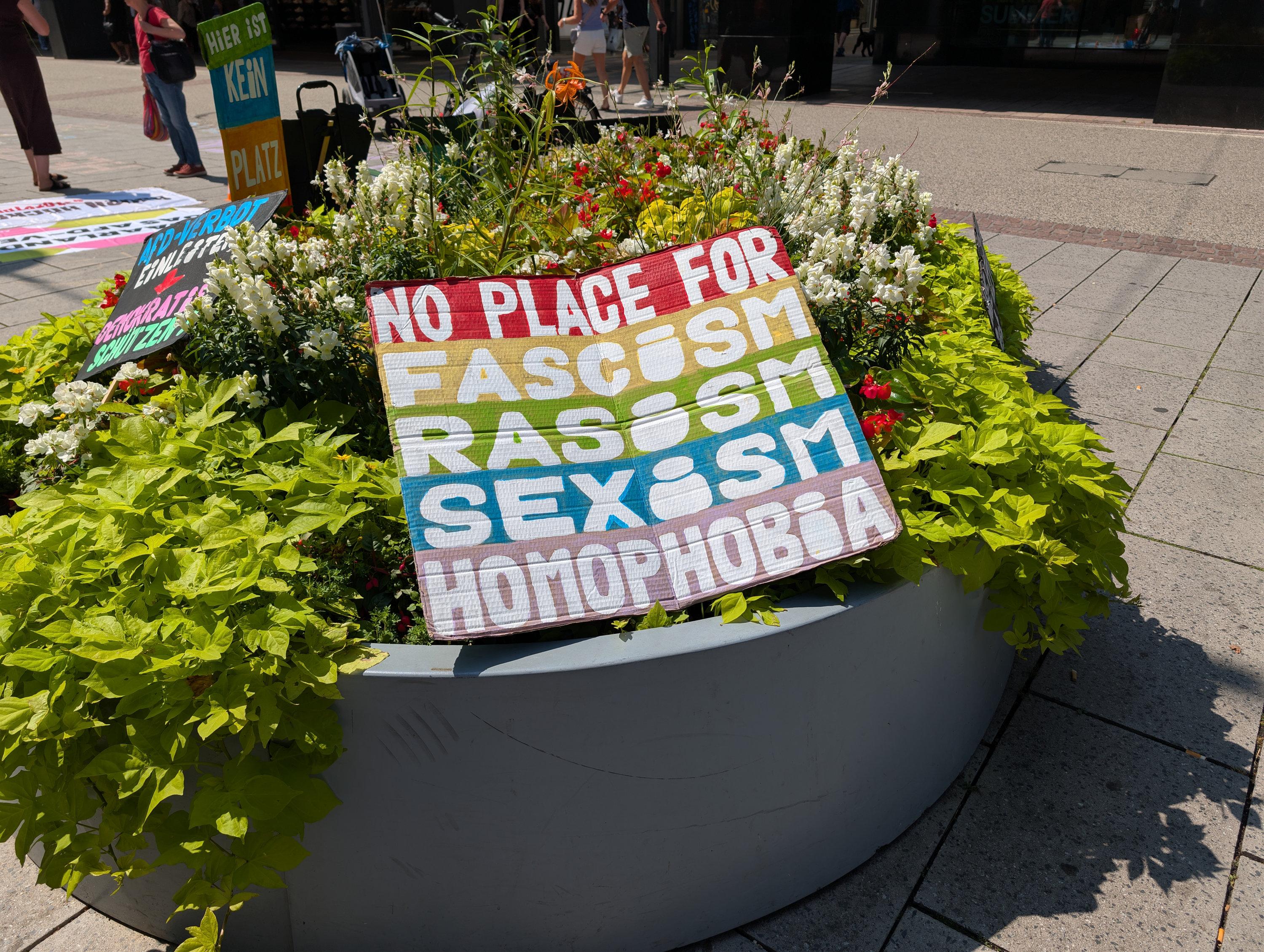 Schild in einem städtischen Blumenarrangement mit dem Aufdruck: No place for fashism, rasism, sexism, Homophobia.