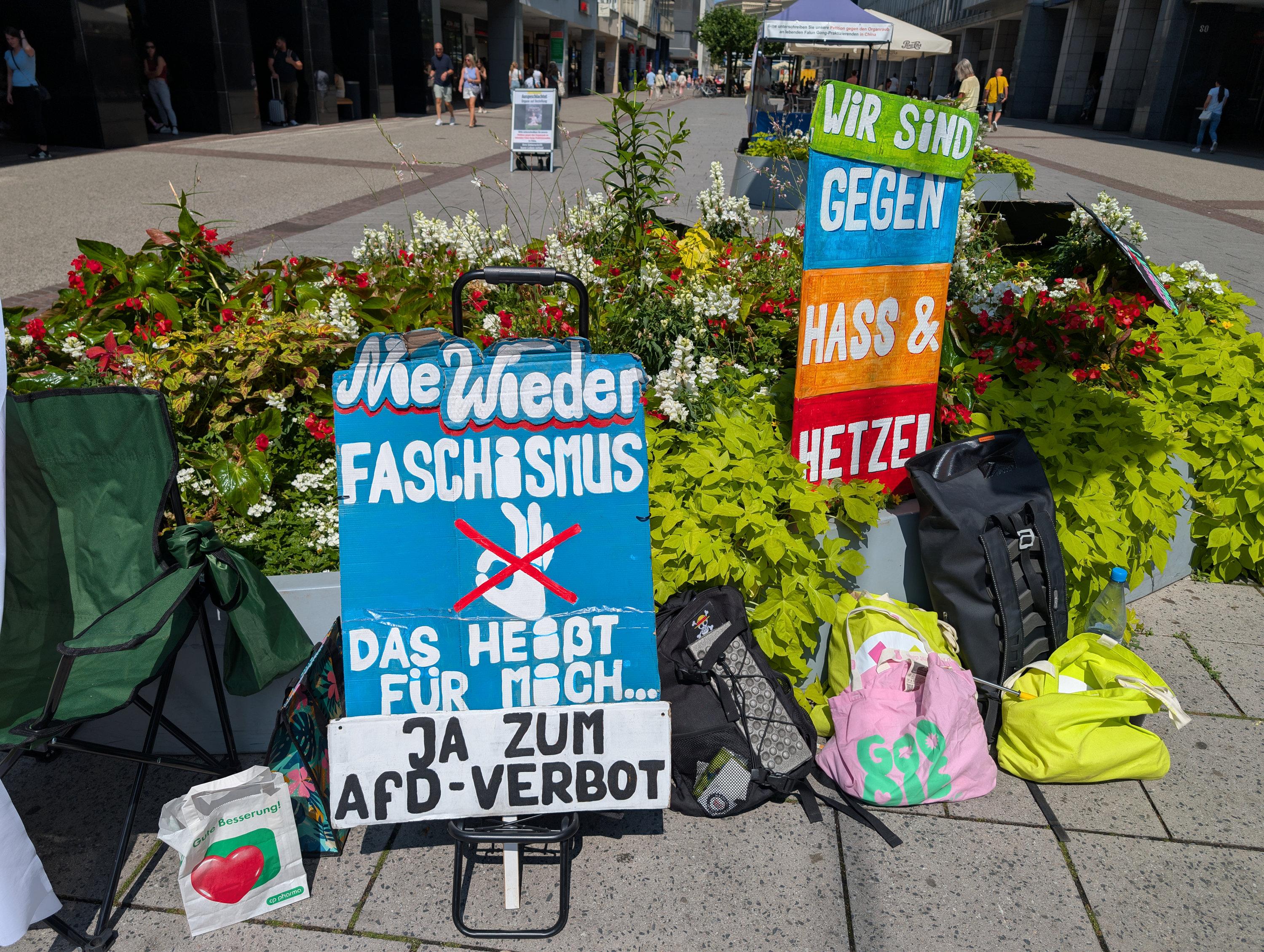 Zwei Schilder in einem städtischen Blumenarrangement mit dem Aufdruck: 1. Wir sind gegen Hass und Hetze. 2. Nie wieder Faschismus. Das heißt für mich Ja zum AfD-Verbot.