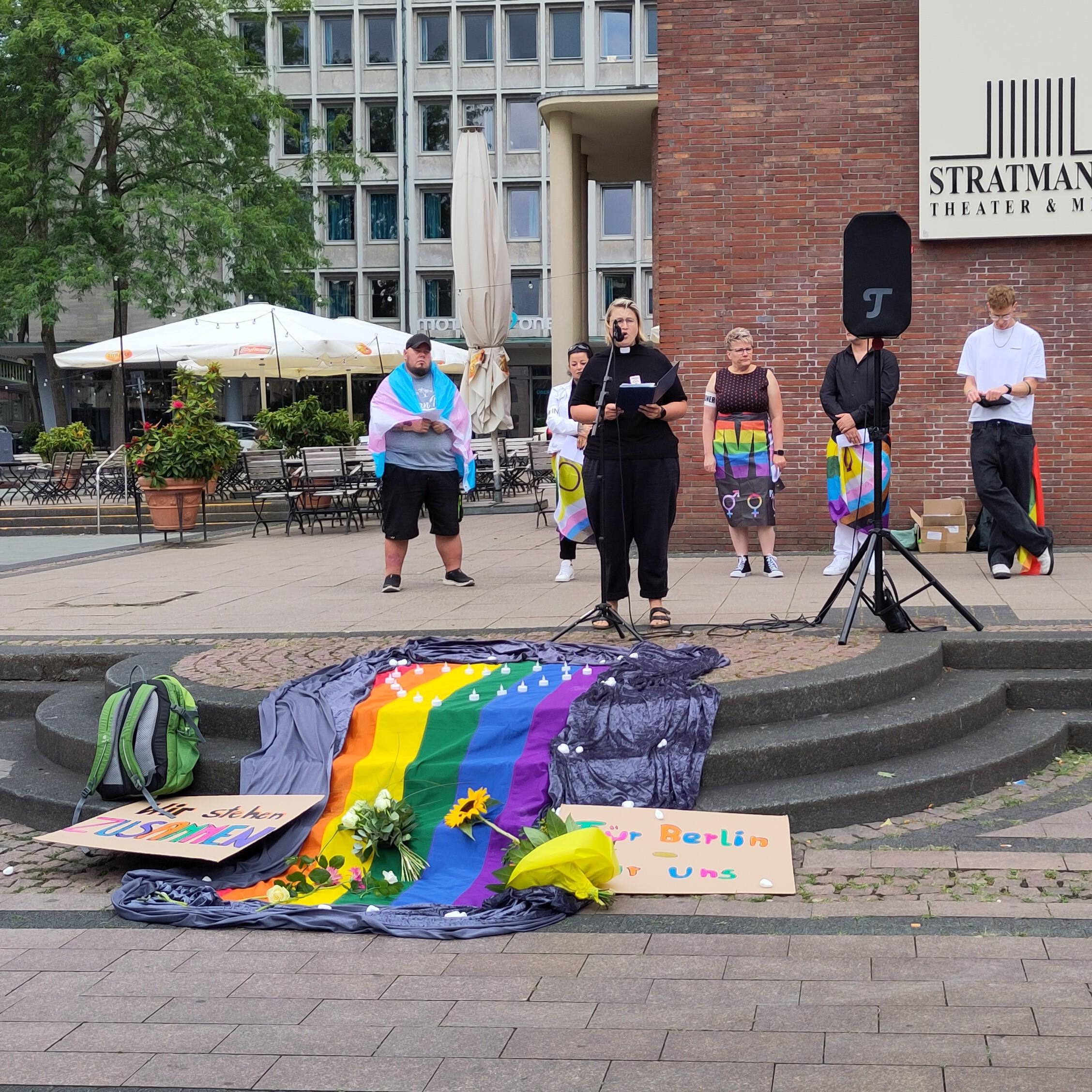 Eine bunte Prideflag auf den Stufen, darauf Blumen. Dahinter ein Mikrofon und 6 Redner*innen
