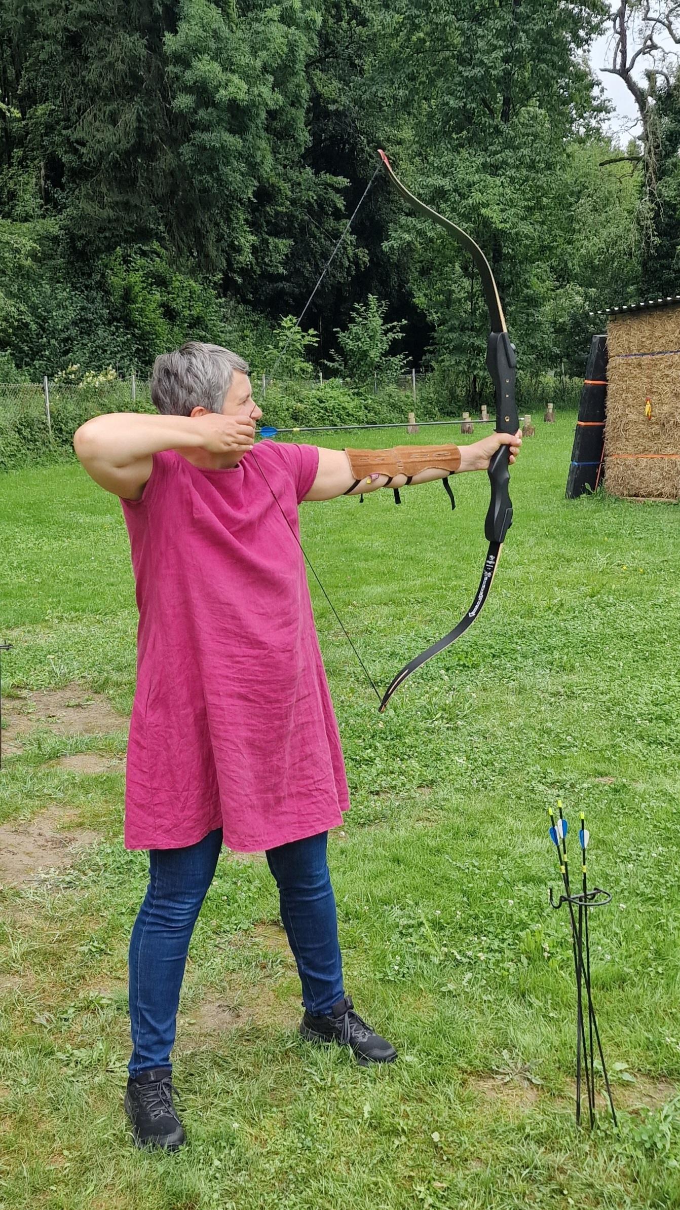 Frau mit kurzem grauem Haar mit pinker Tunika und Bluejeans steht auf einer grünen Wiese und spannt einen Recurve-Bogen zum Schuss auf eine Heuballen-Scheibe im Hintergrund.